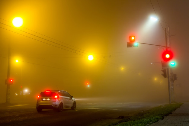 Thick fog over empty road with lonely car and traffic lights at